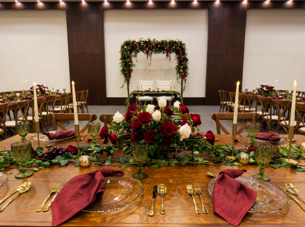 screenshot 20260609 140059 be all inclusive Wedding reception tableside setup with burgundy napkins, gold cutlery, candles, and a lush red-and-white floral centerpiece under a greenery-arched backdrop.