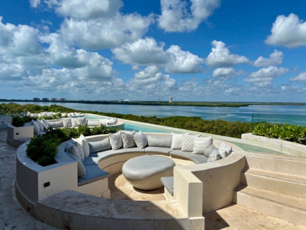 screenshot 20260609 140158 be all inclusive Circular built-in seating with white cushions on a terrace, overlooking a turquoise ocean and blue sky dotted with clouds.