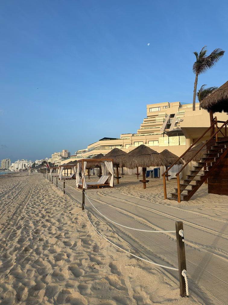 screenshot 20260609 141245 be all inclusive Beach with thatched cabanas and a palm tree along a sandy shore under a clear blue sky. A resort rises in the background to the right.