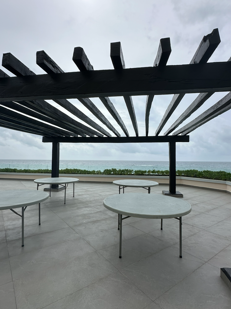 screenshot 20260609 141258 be all inclusive Outdoor terrace with round white tables under a dark wooden pergola, overlooking the ocean on a cloudy day.