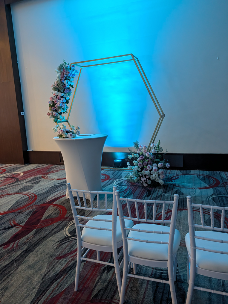 screenshot 20260609 141312 be all inclusive White high-top pedestal table with pastel flower arrangement and a gold hexagonal frame backdrop under blue lighting, with white chairs nearby for a ceremony setting.