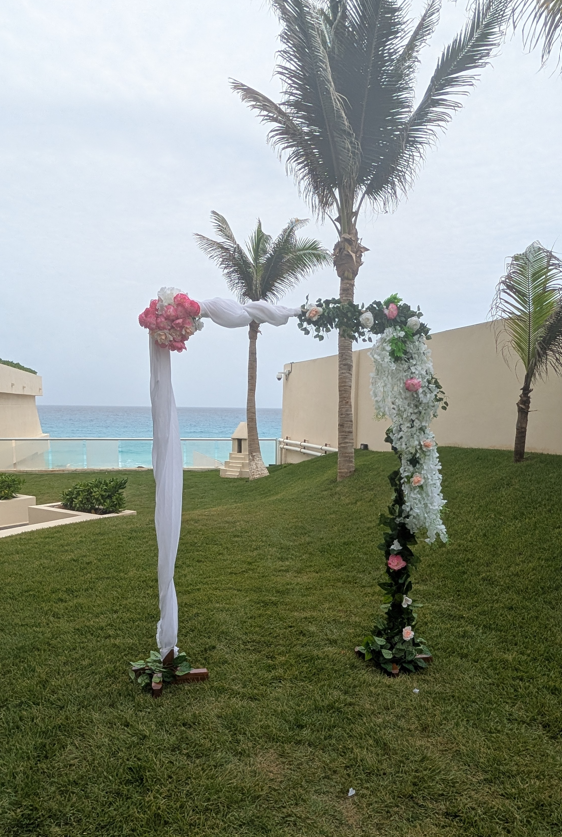 screenshot 20260609 141323 be all inclusive Outdoor wedding arch made of two vertical posts wrapped in white fabric and floral garlands, set on a grassy lawn with palm trees and ocean in the background