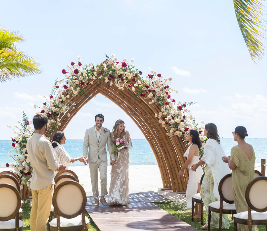 screenshot 20260616 140212 be all inclusive Couple in wedding attire walks under a floral arch on the beach as guests congratulate them.