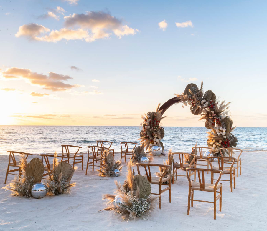screenshot 20260616 140355 be all inclusive Beach wedding ceremony setup at sunset with wooden chairs arranged around a floral arch on white sand.