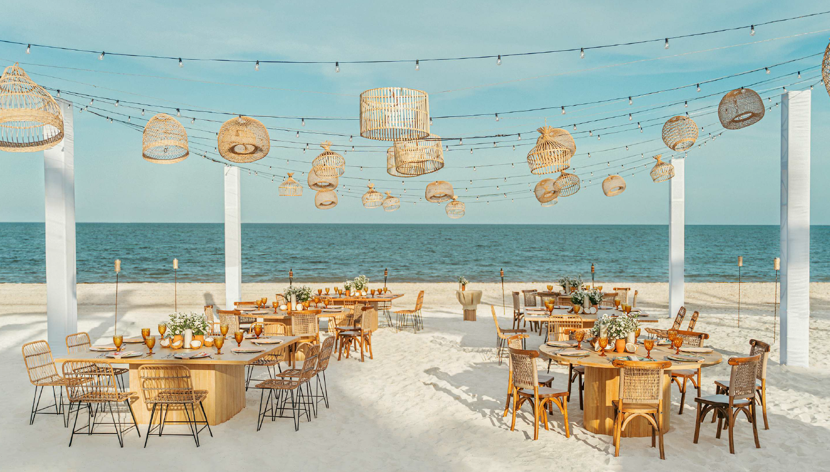 screenshot 20260616 140419 be all inclusive Beachside outdoor dining setup with round and rectangular wooden tables, woven chairs, and string lights over the sand the sea in the distance