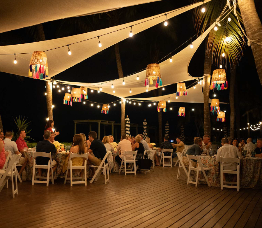 screenshot 20260616 140703 be all inclusive Outdoor evening dinner party under string lights, guests seated at round tables on a wooden deck.