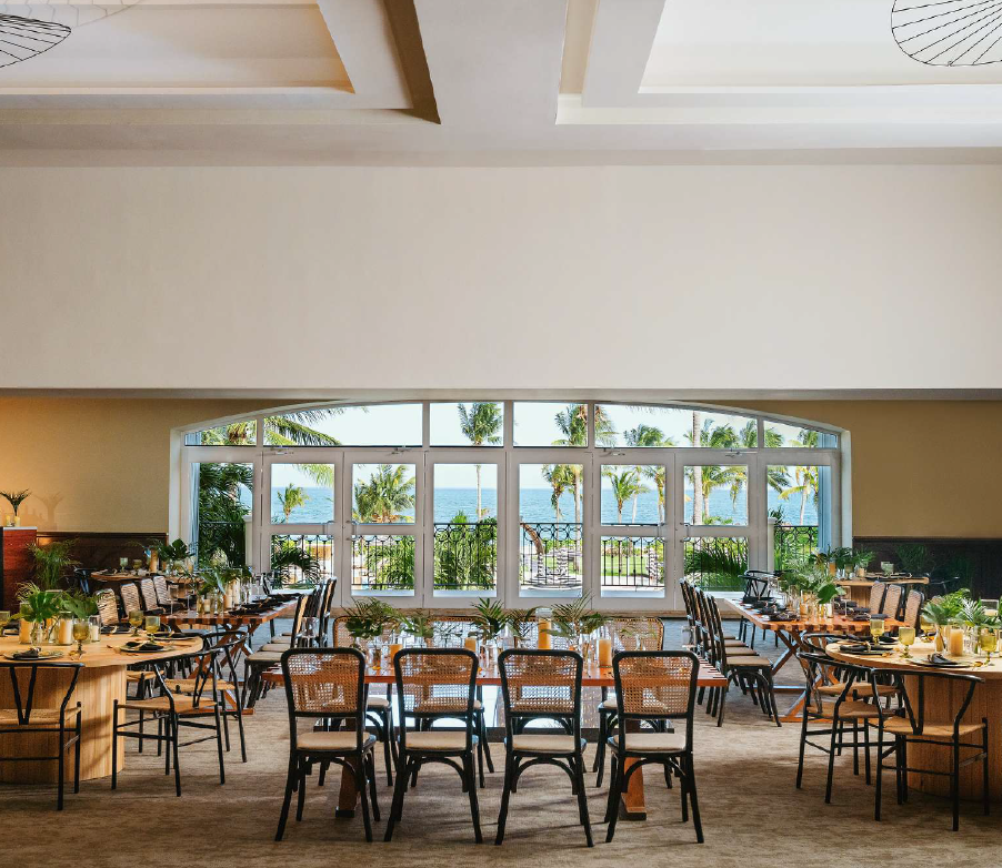screenshot 20260616 141828 be all inclusive Seaside banquet hall set for a meal with long tables and wicker chairs, palm trees and the ocean visible through large glass doors at the back.