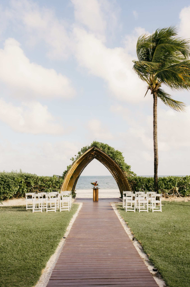 screenshot 20260616 142012 be all inclusive Beach wedding setup with a wooden arched altar, white chairs along a central aisle, and a palm tree nearby.