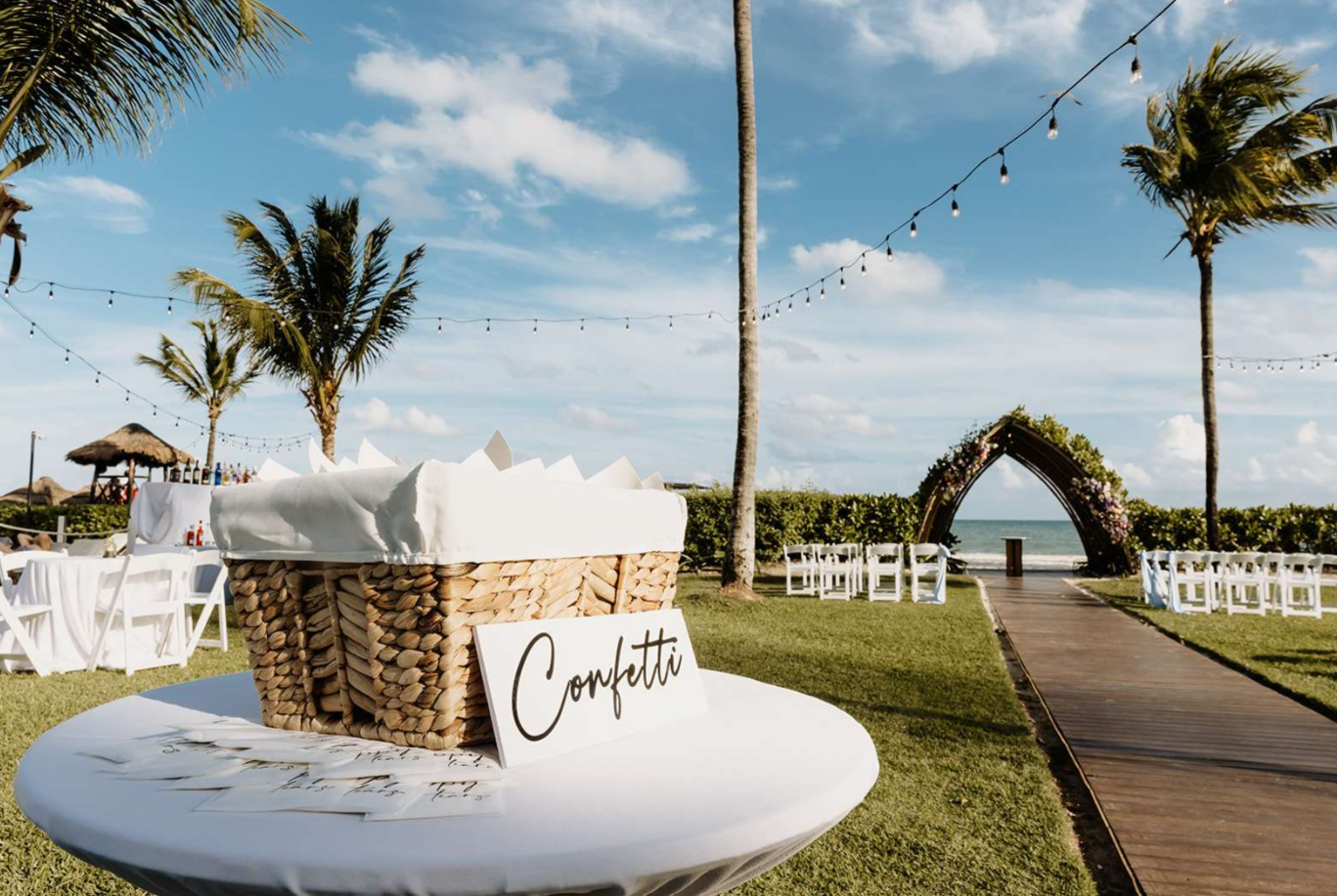 screenshot 20260616 142204 be all inclusive Beach wedding setup along the shore with a flower-decorated wooden arch, string lights, and rows of white chairs facing the ocean; a round table with a woven basket and a 'Confetti' sign in the foreground.