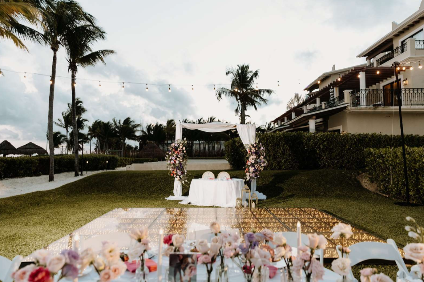 screenshot 20260616 142233 be all inclusive Outdoor wedding ceremony setup with a white draped arch and floral arrangements on a glass dance floor under string lights near a seaside lawn.