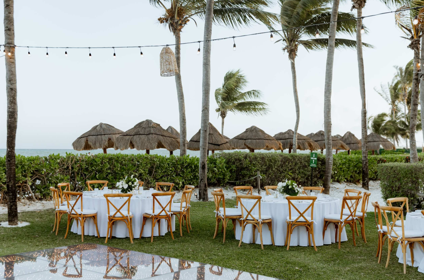 screenshot 20260616 142252 be all inclusive Outdoor beachside event setup with round white‑linen tables, wooden cross‑back chairs, and palm trees under string lights near the ocean edge.