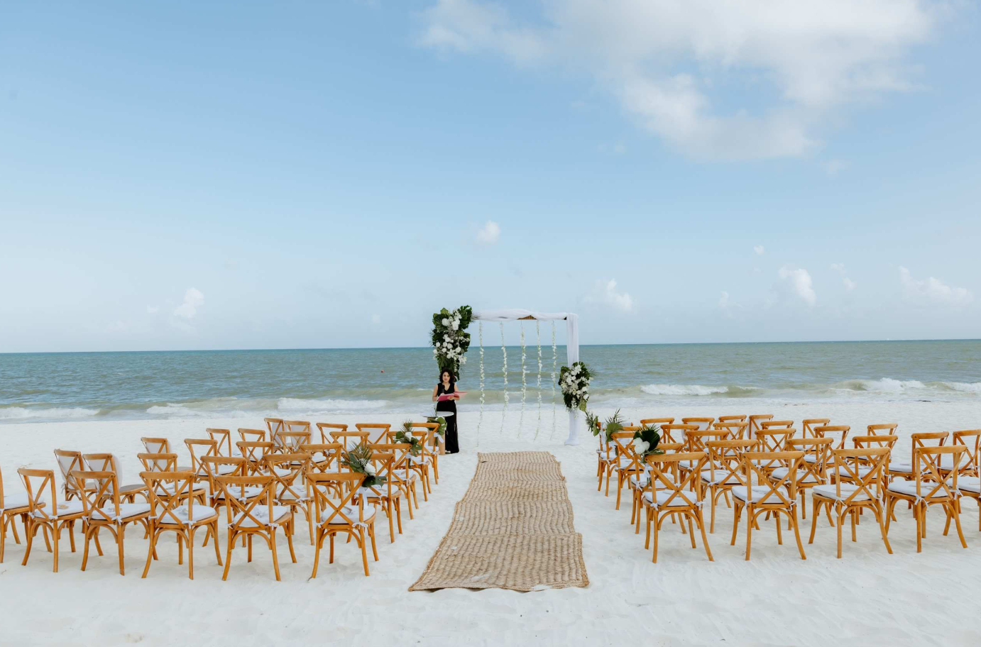screenshot 20260616 142310 be all inclusive Beach wedding setup with wooden chairs facing an arch decorated with white flowers, on white sand by the ocean.