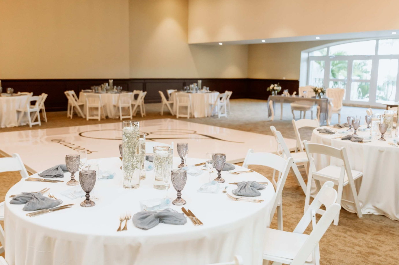 screenshot 20260616 142337 be all inclusive Banquet hall with round white-draped tables, gray napkins, and glass goblets ready for a formal event, centerpieces in tall glass vases start the décor.
