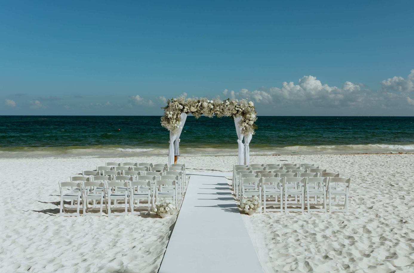 screenshot 20260616 142353 be all inclusive White wedding arch covered in white flowers on a beach, with rows of white chairs and a white aisle runner leading to the sea.