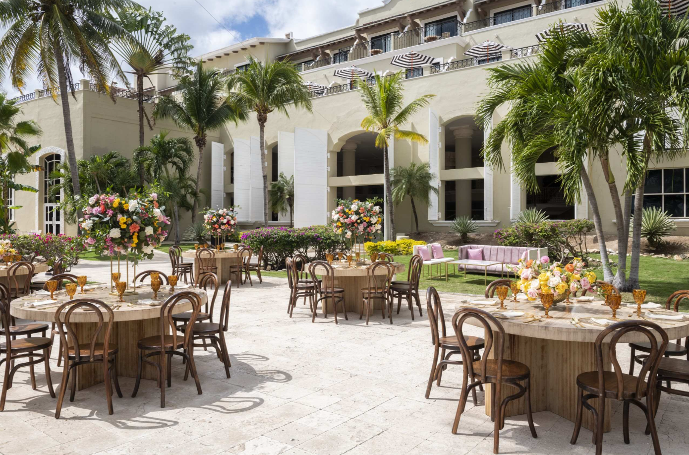 screenshot 20260616 143008 be all inclusive Outdoor wedding reception setup on a sunny patio with round wooden tables, floral centerpieces, and gold-glass tableware under palm trees by a beige hotel building