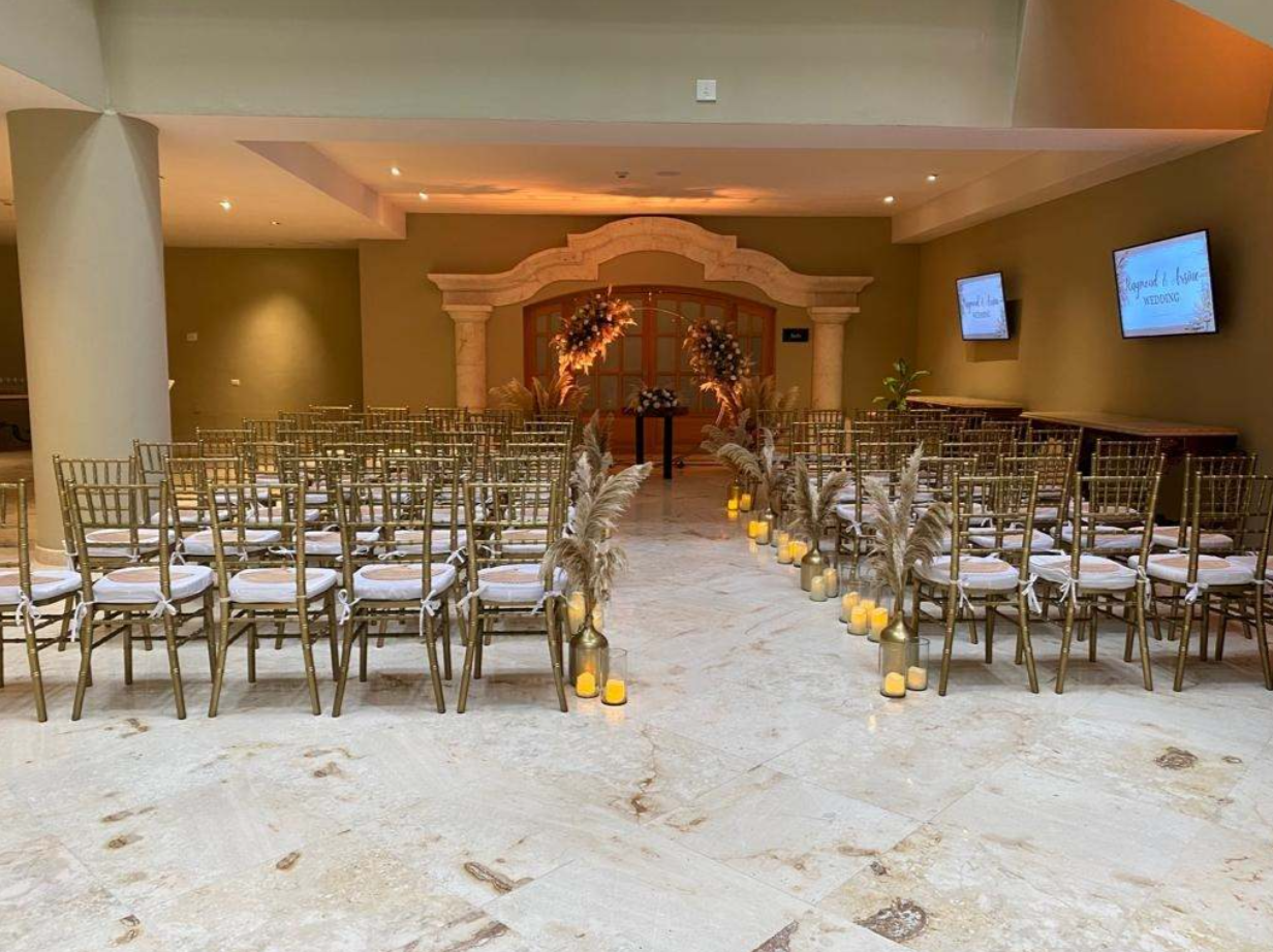 screenshot 20260616 143038 be all inclusive Wedding ceremony setup: two rows of gold chairs facing a floral arch at the front, with a lit candle aisle lined by vases and pampas grass.