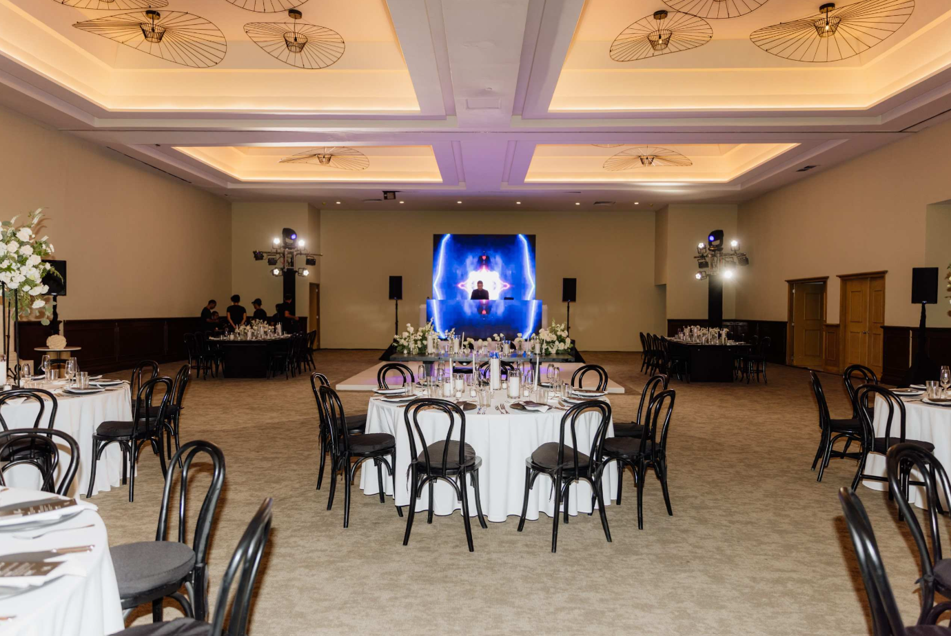 screenshot 20260616 143118 be all inclusive Banquet hall set for a formal event with round tables, white linens, black chairs, and a blue-lit DJ stage in the background.
