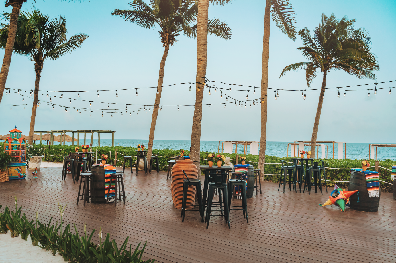 screenshot 20260616 143221 be all inclusive Beachside outdoor cafe on a wooden deck with string lights, palm trees, and the ocean in the distance, tables set with flowers.