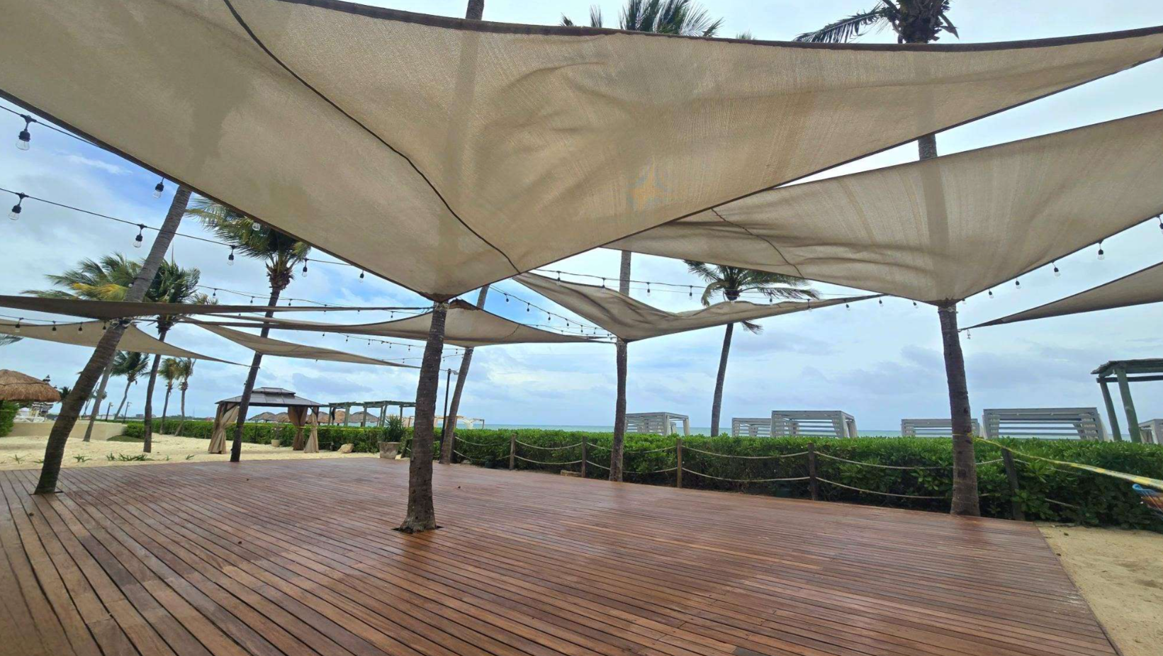 screenshot 20260616 143238 be all inclusive Wooden beach deck with palm trees and large fabric shade sails, string lights, and a view of the sea in the distance.