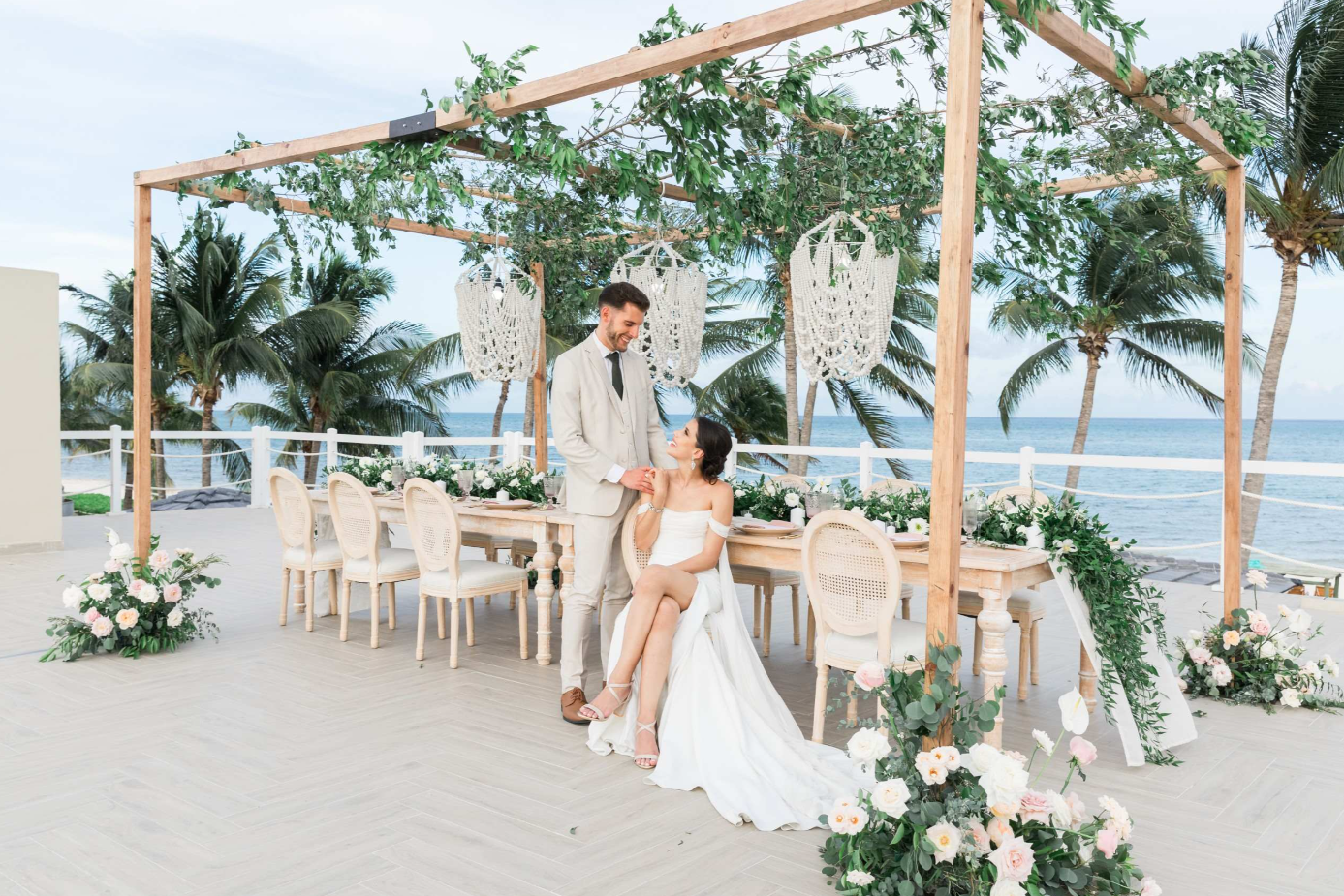 screenshot 20260616 143259 be all inclusive Bride in a white gown sits on a chair under a wooden arch with the groom standing beside her on a beachside wedding setup; ocean in the background and floral decorations nearby.