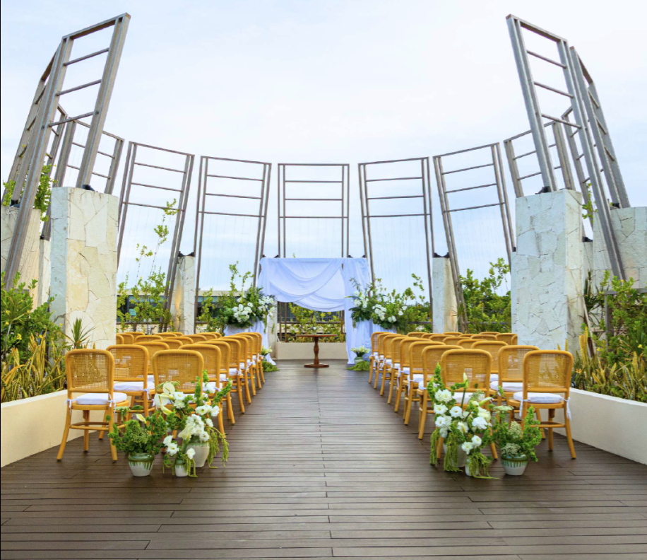 screenshot 20260630 123834 be all inclusive Outdoor wedding ceremony setup with wooden aisle, yellow chairs, and white floral arrangements facing an altar under a curved metal framework.
