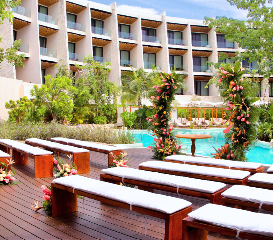 screenshot 20260630 123939 be all inclusive Resort pool area with white-cushioned wooden lounge benches, floral arches, and a multi-story building in the background.