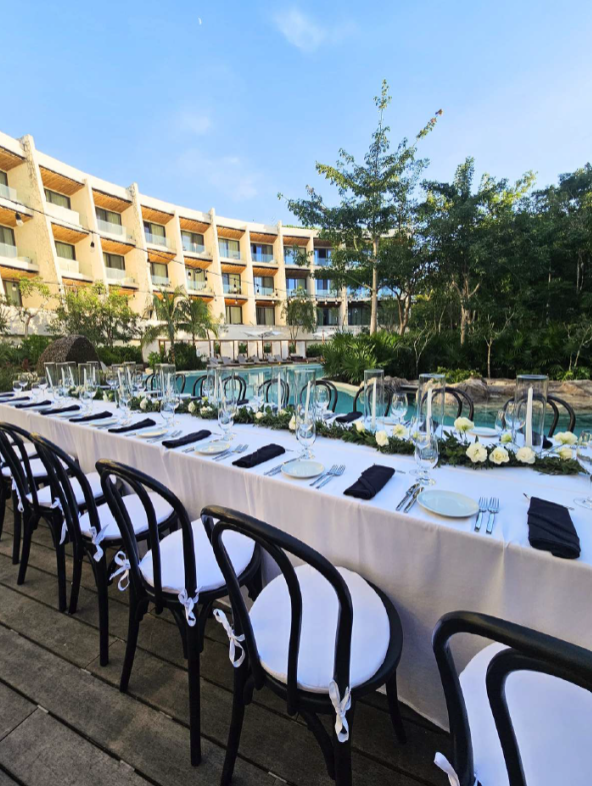 screenshot 20260630 124012 be all inclusive Long outdoor poolside dining table set with white tablecloth, black napkins, glassware, and white flowers, with hotel balconies in the background.