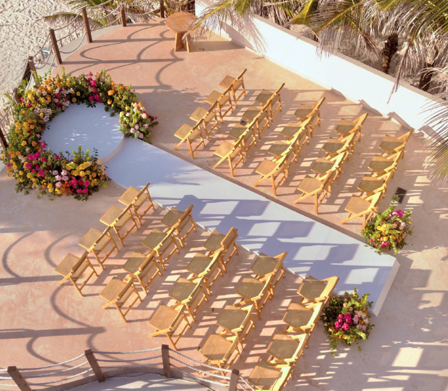 screenshot 20260630 124124 be all inclusive Outdoor wedding ceremony setup with wooden chairs arranged in rows around a white aisle, flanked by colorful flower arches and tropical plants.