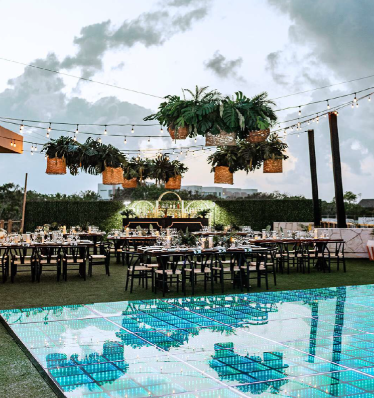 screenshot 20260630 124357 be all inclusive Outdoor evening wedding reception with hanging planters, string lights, and neatly arranged round tables on the lawn near a glass-bottom pool reflection.