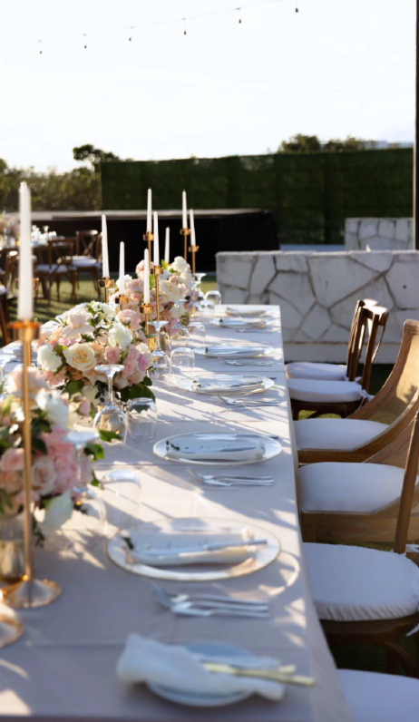 screenshot 20260630 124452 be all inclusive Outdoor long banquet table with white cloth, floral centerpieces, gold candlesticks, and neatly arranged plates and cutlery on a sunlit terrace.”
