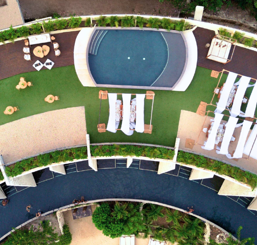 screenshot 20260630 124643 be all inclusive Aerial view of a resort pool area with a circular pool, white cabanas and loungers, wooden decks, and curved green pathways surrounded by plants.