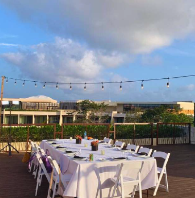 screenshot 20260630 124807 be all inclusive Outdoor rooftop dinner setup: long white-cloth table with place settings and centerpieces, white folding chairs, string lights, under a blue sky with buildings in the distance.