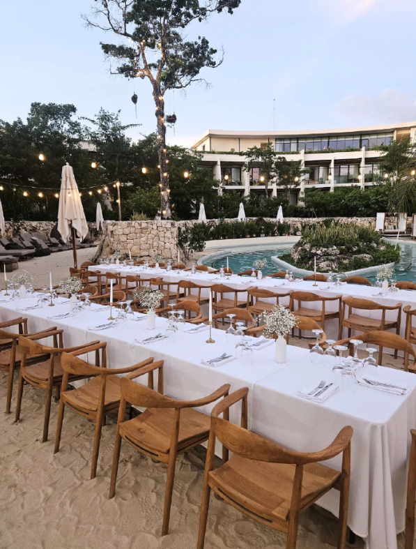 screenshot 20260630 124939 be all inclusive Outdoor beachside dining setup with a long white-clothed table, wooden chairs, and floral centerpieces under string lights near a pool and resort building.