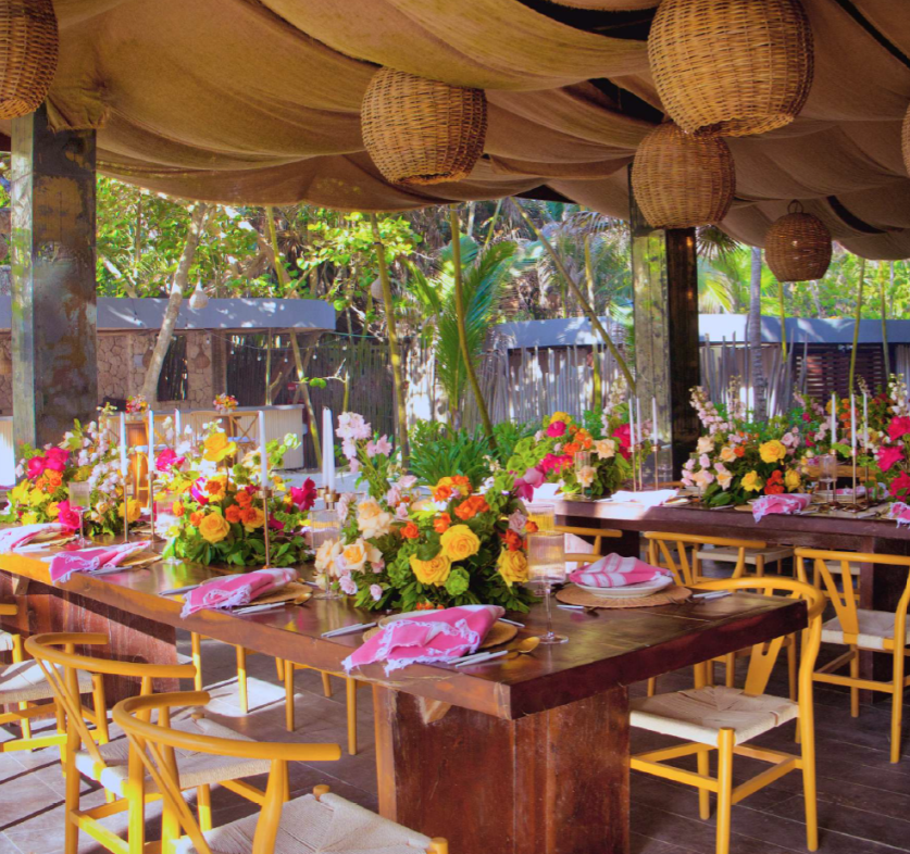 screenshot 20260630 125241 be all inclusive Outdoor event setup with long wooden tables, colorful flower centerpieces, candles, and wicker lanterns under a draped canopy.