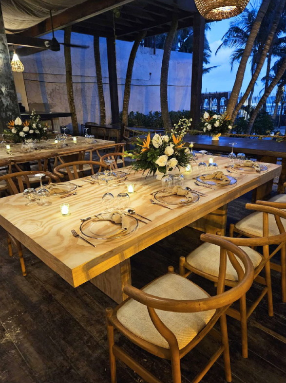 screenshot 20260630 125307 be all inclusive Long wooden dining table set for a formal outdoor dinner with floral centerpiece, candles, and place settings under a covered patio.