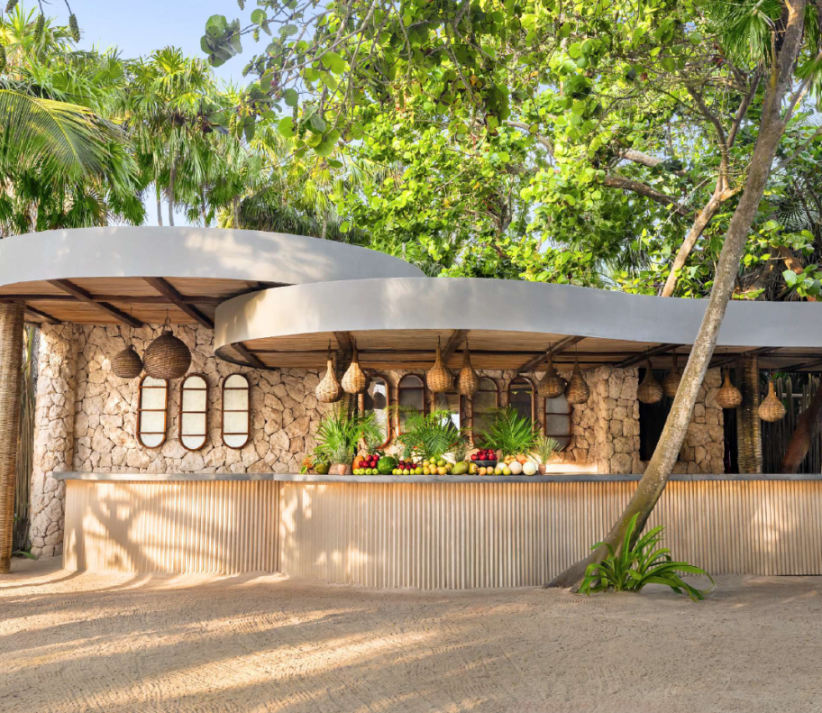 screenshot 20260630 125444 be all inclusive Tropical open‑air bar with stone walls, curved white canopy, and a colorful fruit display on the counter under hanging baskets.