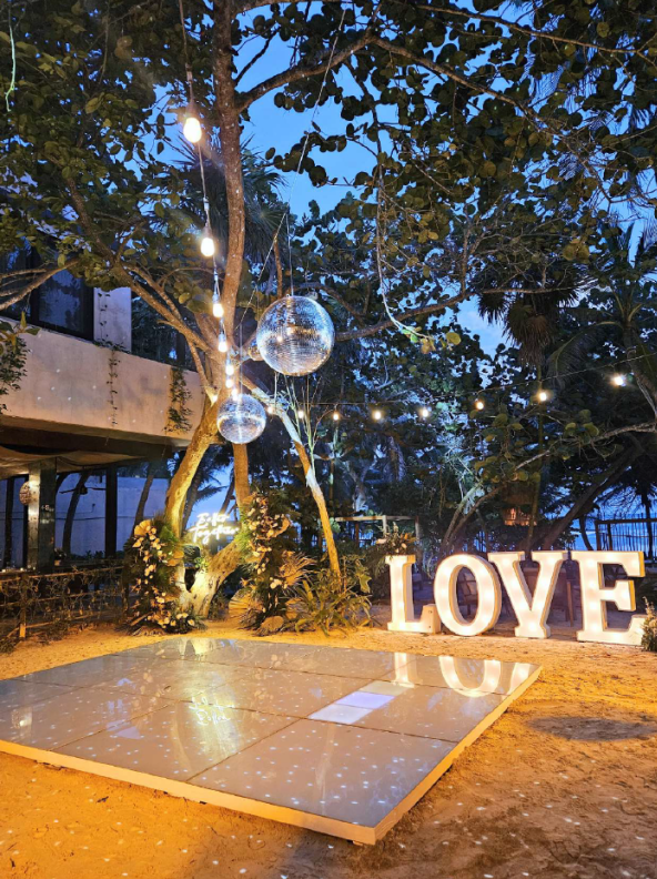 screenshot 20260630 125508 be all inclusive Outdoor evening scene with string lights, a hanging disco ball, and large illuminated letters spelling LOVE beside a small reflective dance floor.