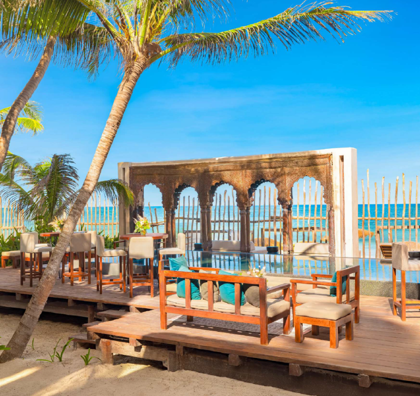 screenshot 20260630 125643 be all inclusive Beachside resort seating on a wooden deck with couches, palm trees, and a decorative arch by a pool overlooking the ocean
