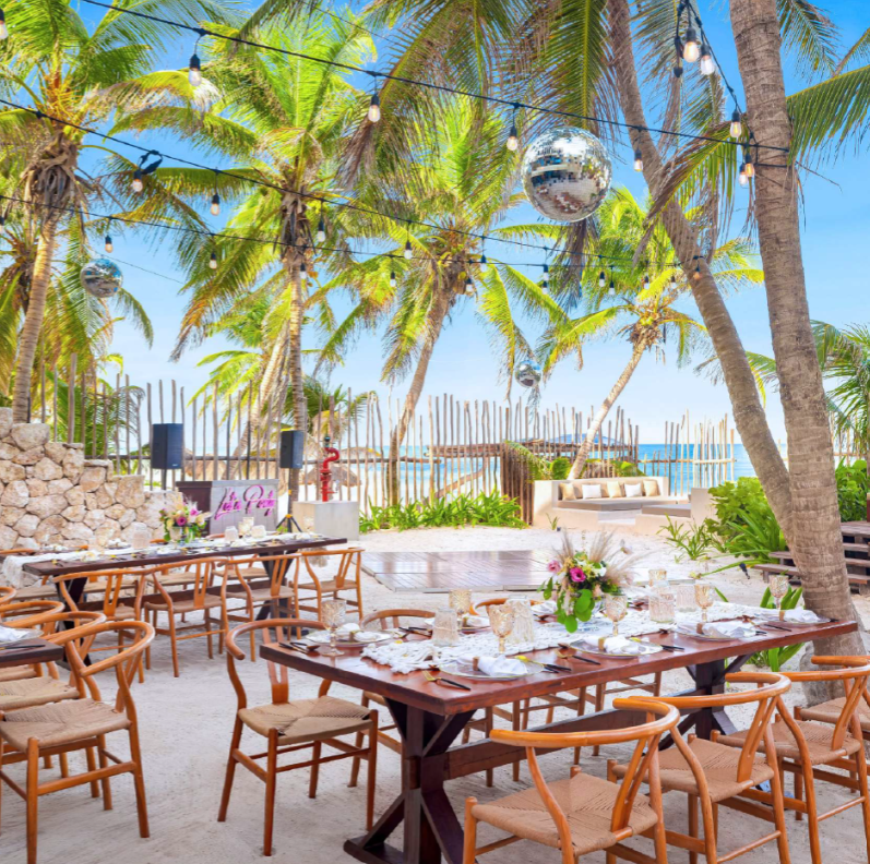 screenshot 20260630 125712 be all inclusive Outdoor tropical beachside dining setup with wooden tables, chairs, string lights, and a disco ball among palm trees over a sandy venue by the sea.