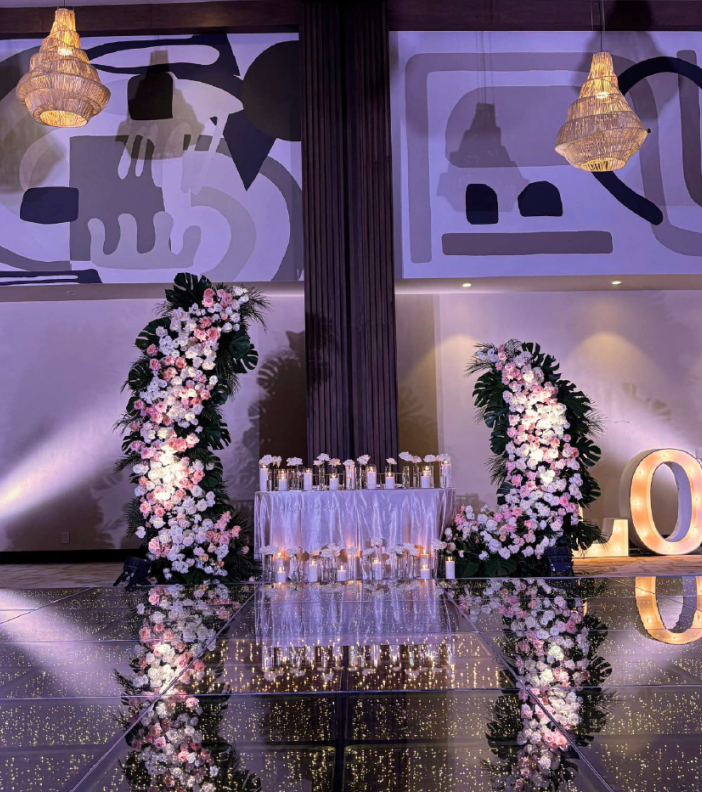 screenshot 20260630 132437 be all inclusive Elegant event stage with two tall floral arches framing a table of champagne flutes on a mirrored, glittering floor; illuminated LO letters on the right.