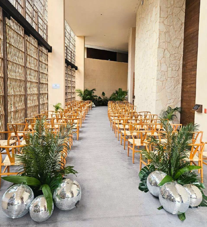 screenshot 20260630 132606 be all inclusive Rows of light wood chairs arranged along a central aisle in a modern indoor courtyard, with decorative silver spheres and green plants at the front.