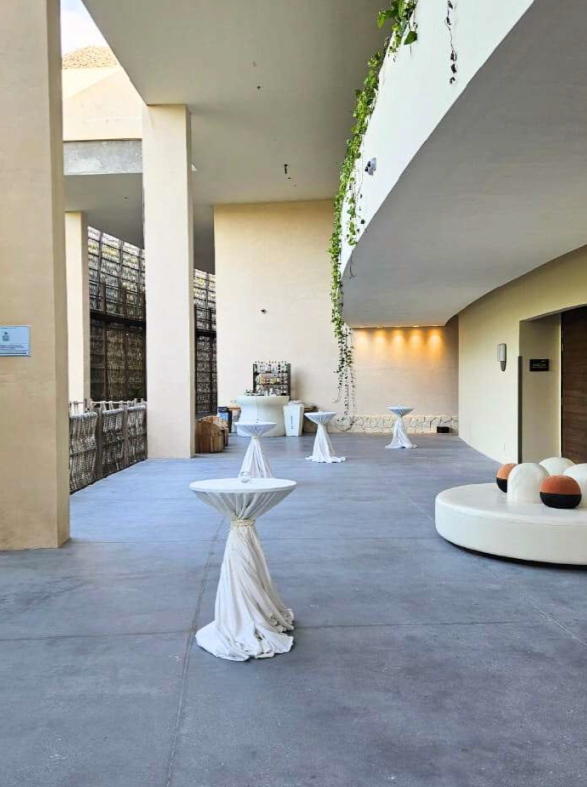 screenshot 20260630 132623 be all inclusive Open-air courtyard with tall beige columns, cocktail tables draped in white cloths, and a circular seating area with orange cushions.