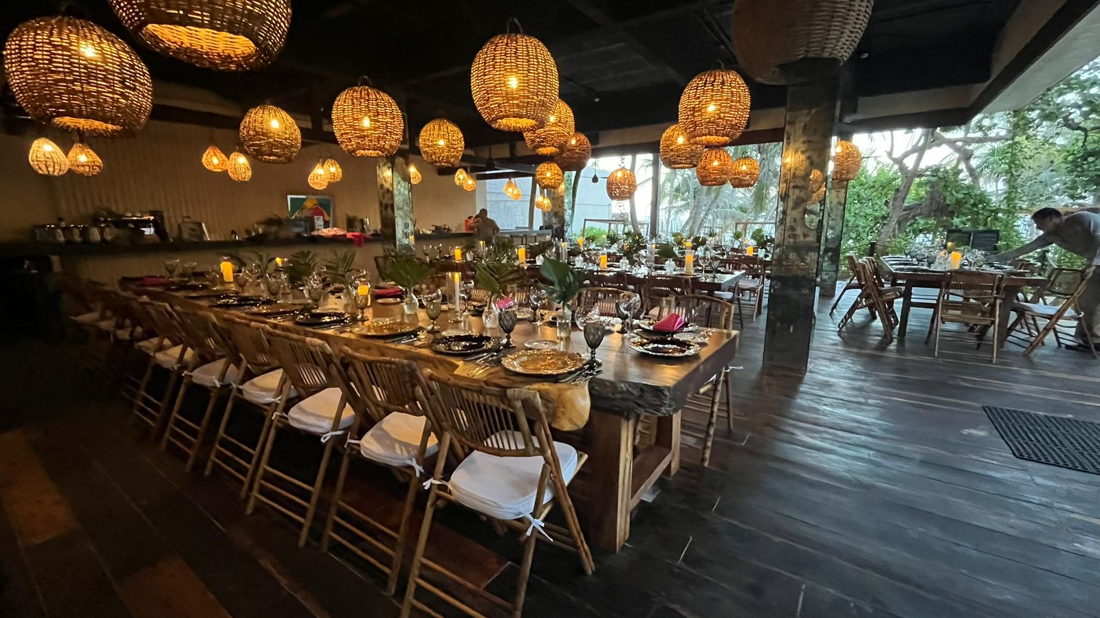 seaside 1 be all inclusive Long wooden table set for a formal dinner with woven pendant lights and candles in a tropical-style restaurant interior with greenery outside.