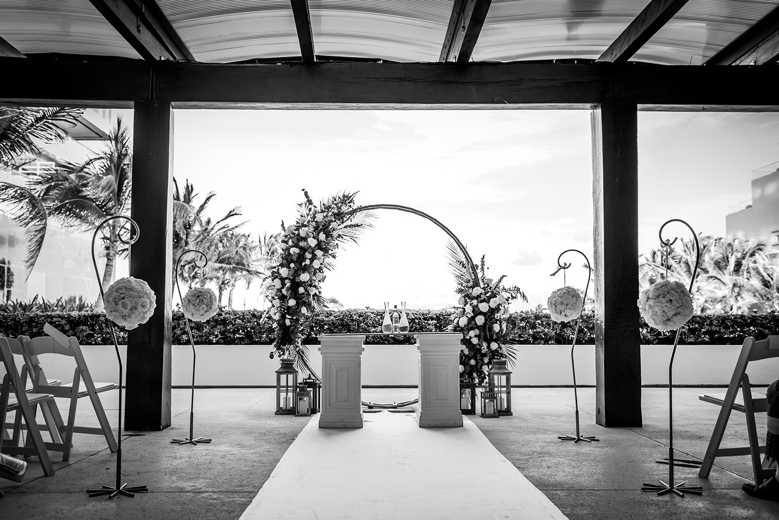 secrets the vine be all inclusive Wedding ceremony setup with a white aisle runner leading to a floral arch and two white pillars, flanked by decorative stands and lanterns.