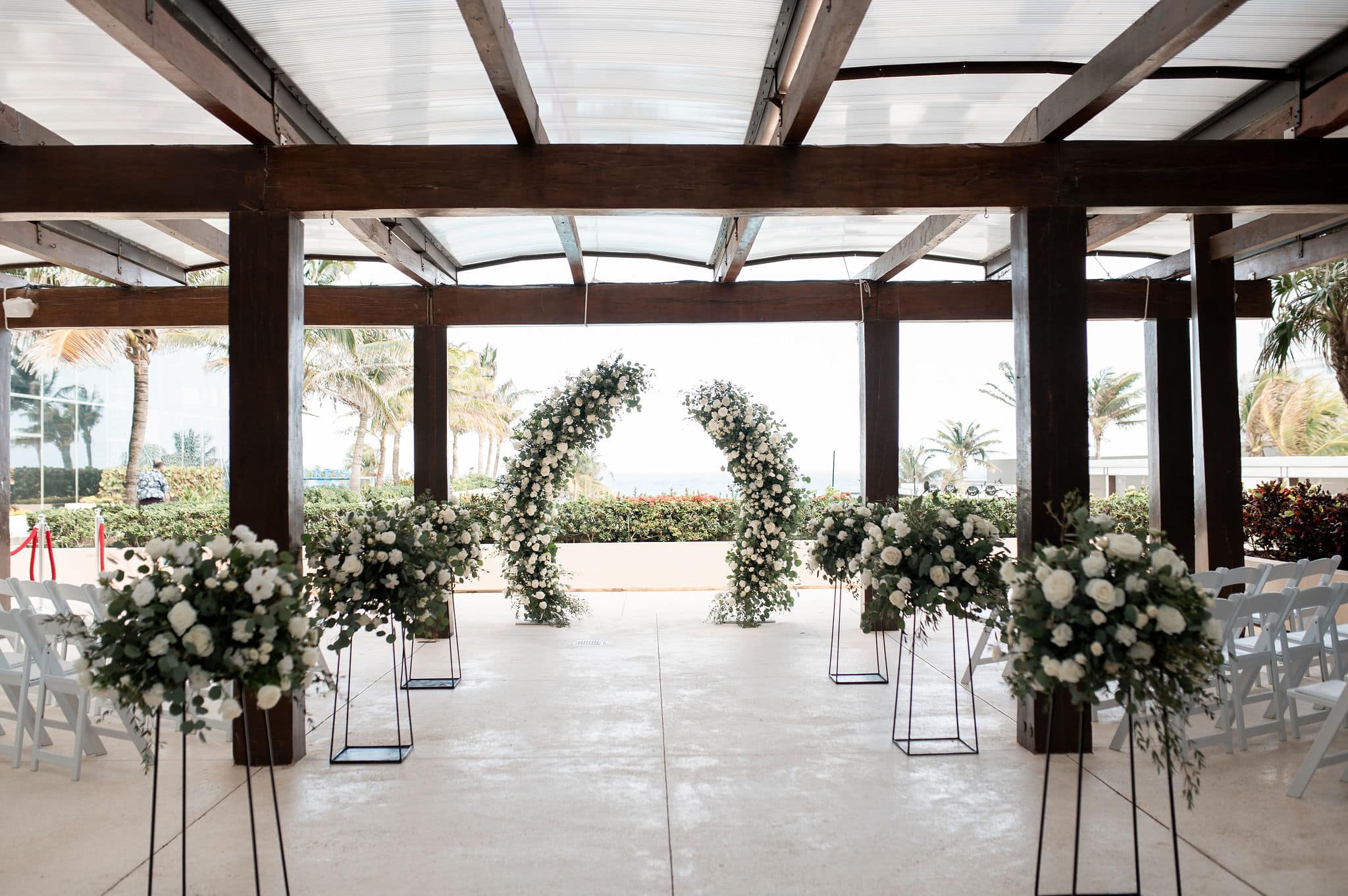 tlvt 5 be all inclusive Outdoor wedding ceremony setup with white floral arches, wooden beams, and white folding chairs facing an aisle.