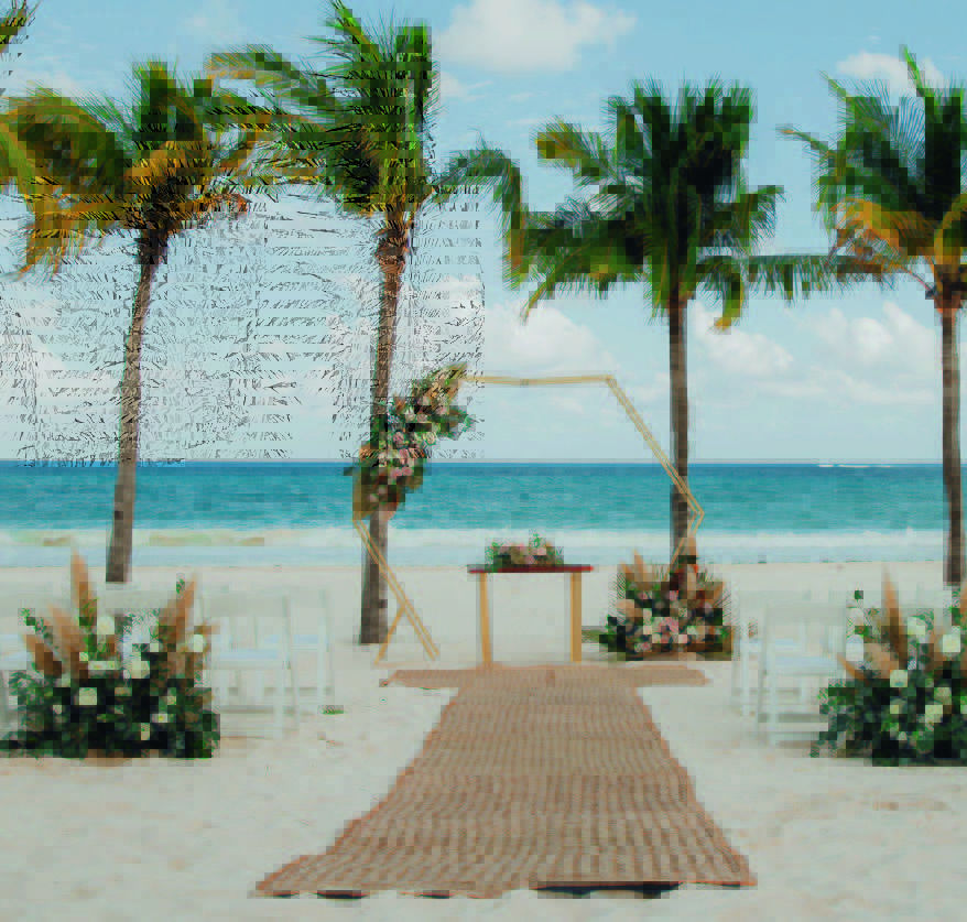 wedding guide 2026 be all inclusive Beach wedding setup: floral arch over a woven aisle, wooden table with flowers, palm trees and ocean backdrop on white sand.