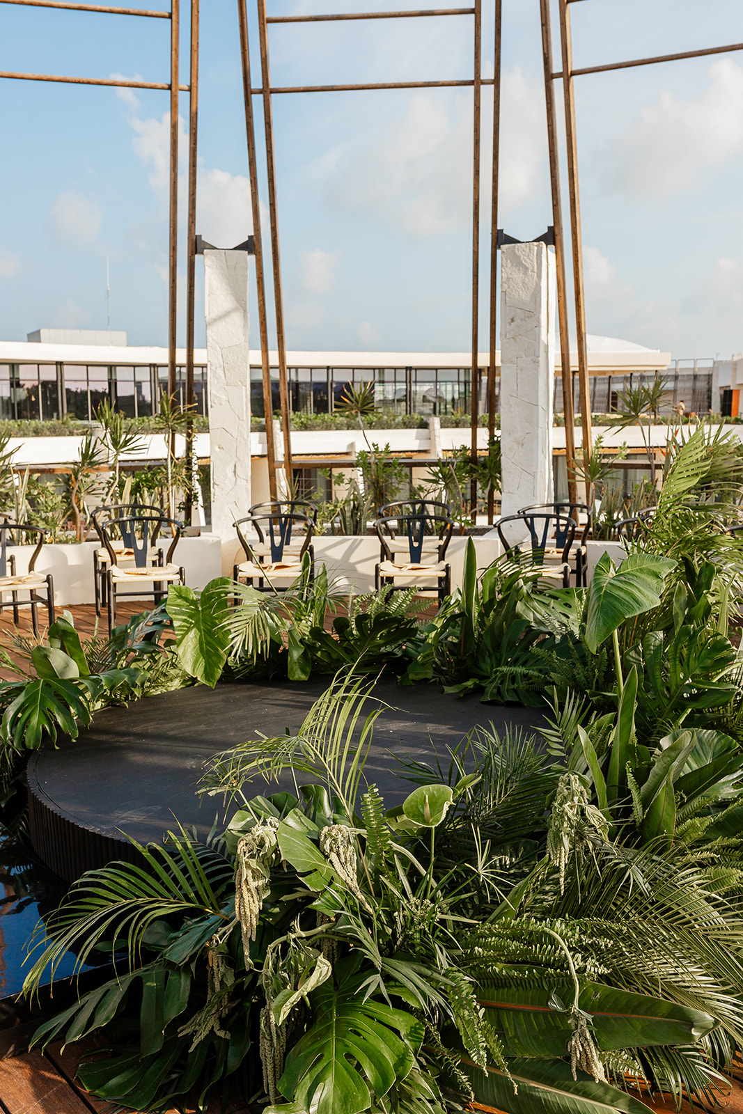 waylon zack be all inclusive Outdoor terrace with lush tropical plants surrounding a circular black stage and metal framework, seating area in the background.