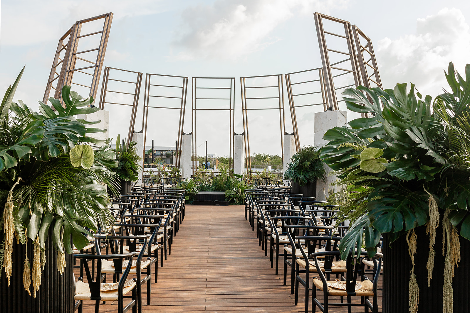 waylon zack be all inclusive Outdoor ceremony space with a wooden aisle, black chairs, and lush tropical plants along the sides beneath tall wooden arch frames.