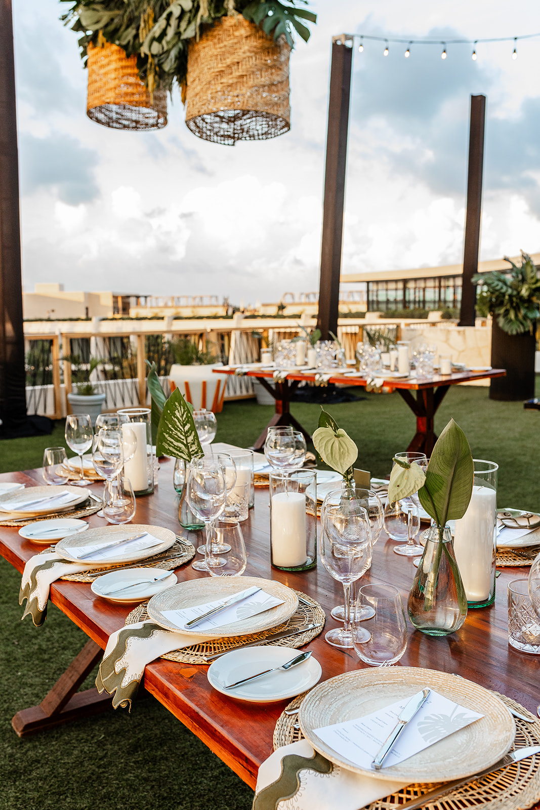 waylon zack be all inclusive Outdoor rooftop dining setup with wooden tables, candles, glassware, and tropical leaves under string lights.
