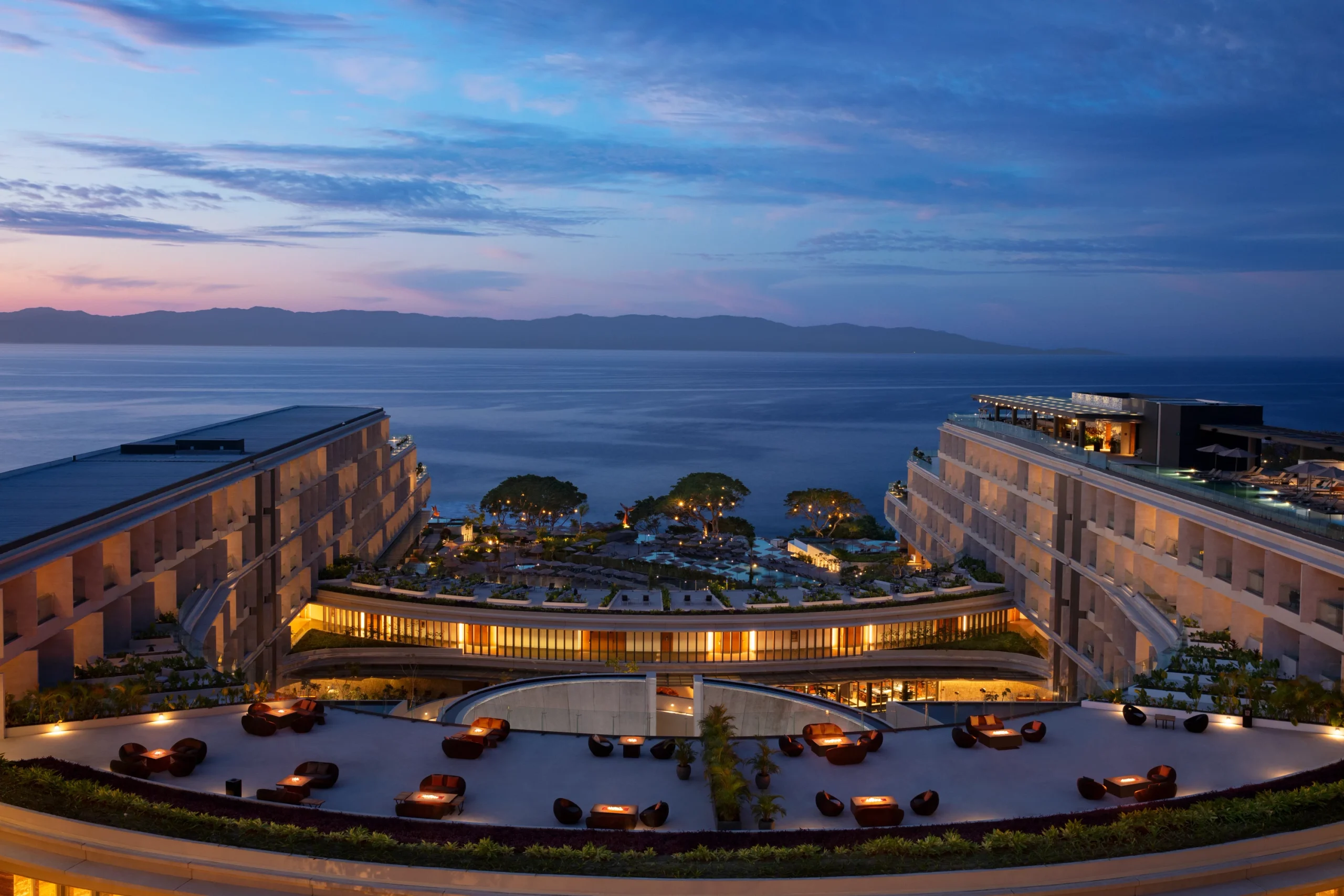 drbmiextmoondeckpano2cbresized6800374f682d3 be all inclusive Resort complex at dusk overlooking calm ocean with distant mountains beyond the water's edge and illuminated courtyards.